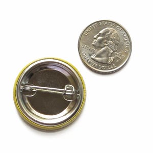 May include: A circular pin-back button with a metallic silver back and a horizontal metal pin mechanism, positioned next to a United States quarter dollar coin for scale, showing the reverse side of the button with its fastening hardware.