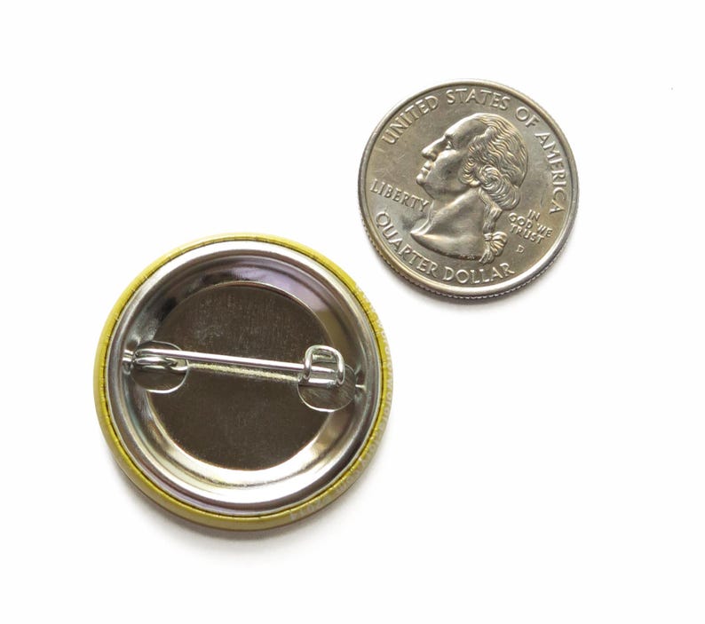 May include: A circular pin-back button with a metallic silver back and a horizontal metal pin mechanism, positioned next to a United States quarter dollar coin for scale, showing the reverse side of the button with its fastening hardware.