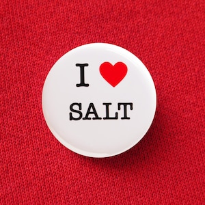 Może przedstawiać: Biała przypinka z napisem "I ❤️ SALT" w kolorze czarnym i czerwonym. Przypinka jest umieszczona na czerwonym tle z tkaniny. Przypinka jest okrągła i ma błyszczącą powierzchnię.
