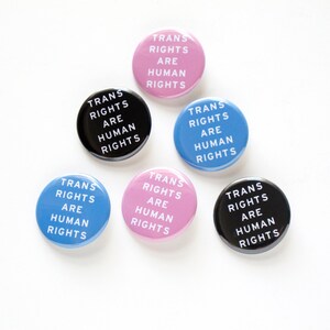 A triangle of 6 round pinback button that read TRANS RIGHTS ARE HUMAN RIGHTS in white text. They are a mix of blue, pink & black background colors.