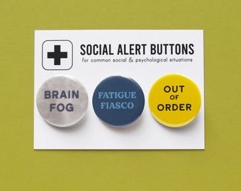 Può includere: Tre bottoni rotondi con frasi diverse. I bottoni sono bianchi, blu e gialli. Il testo sui bottoni recita "Brain Fog", "Fatigue Fiasco" e "Out of Order".