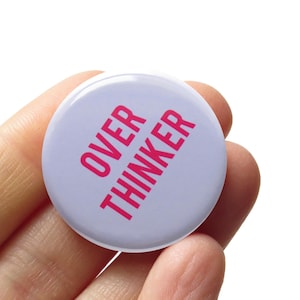 May include: A round pinback button with a light lavender background and the text OVER THINKER printed in bold, bright pink capital letters. The button has a smooth, glossy finish and a circular shape, featuring clear, sans-serif typography.