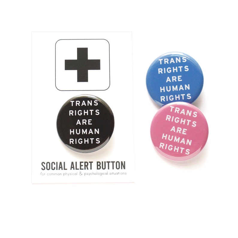 A round black pinback button that reads TRANS RIGHTS ARE HUMAN RIGHTS in white text. Button is on a Social alert Button backing card. Blue & pink versions of the button are to the right.