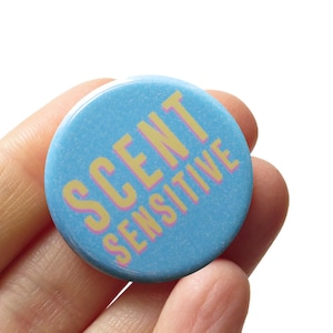 May include: A round, light blue button with the words "SCENT SENSITIVE" in yellow and pink lettering. The button is held in a person's hand against a white background. The button is a wearable accessory.