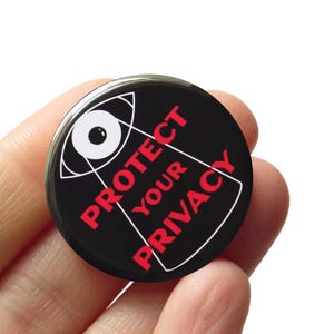 May include: A circular pin-back button with a black background featuring a white graphic of an eye and the red text "PROTECT YOUR PRIVACY" arranged in a triangular shape. The design includes white lines forming a pyramid outline around the text.