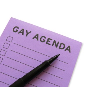 Close up off the top of a vertical rectangle lavender notepad that reads GAY AGENDA at the top, with check boxes & lines for list making. A black felt tip pen is in the foreground.