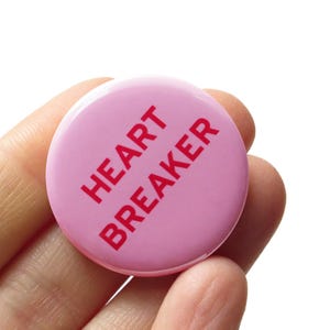 A round light pink pinback button that reads HEART BREAKER' in dark pink text on 2 lines. The button is held in a hand.