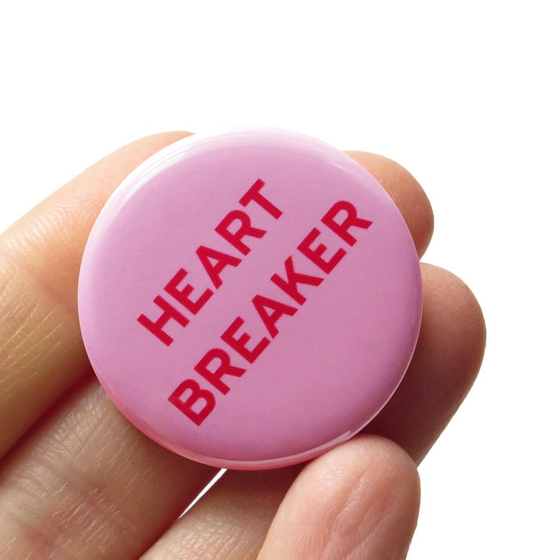 A round light pink pinback button that reads HEART BREAKER' in dark pink text on 2 lines. The button is held in a hand.