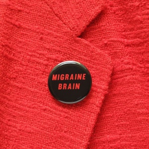 A round black pinback button that reads MIGRAINE BRAIN in red text, pinned to a red woven blazer lapel.