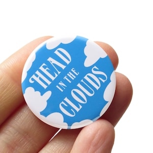 May include: A round, blue button with white clouds and the text "HEAD IN THE CLOUDS" in white, stylized font. The button is held in a hand against a white background. The button is a fun accessory.