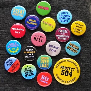 A bunch of colorful round pinback buttons that relate to disability, chronic illness & symptoms with some social justice pins thrown. Badges are pinned to black denim.