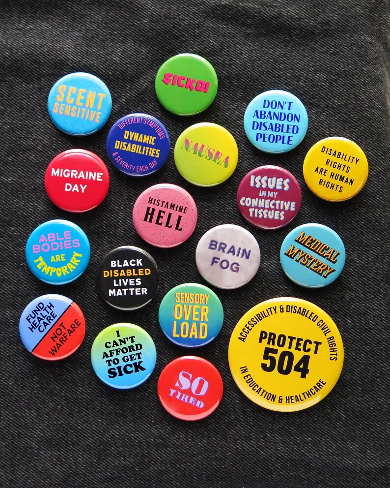 A bunch of colorful round pinback buttons that relate to disability, chronic illness & symptoms with some social justice pins thrown. Badges are pinned to black denim.