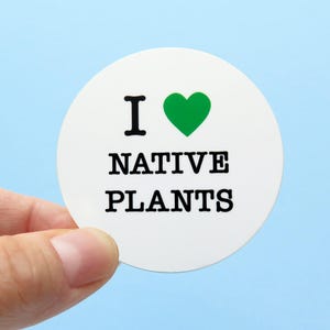 A round white sticker that reads I LOVE NATIVE PLANTS, love being indicated by a green heart. Held in the lower right corner with 2 fingers.