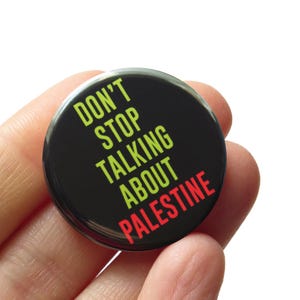 May include: A black button with the text "Don't Stop Talking About Palestine" in yellow and red lettering.
