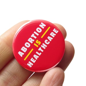 May include: Red button with white text that reads "ABORTION IS HEALTHCARE" with a yellow line through the word "IS".