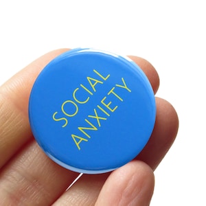 May include: A circular blue pin-back button featuring the text SOCIAL ANXIETY printed in yellow capital letters. The button has a smooth, glossy finish and a simple, clean design with the text oriented vertically across the center of the surface.