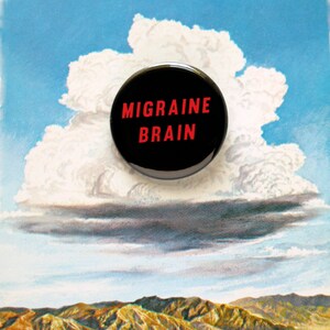 A round black pinback button that reads MIGRAINE BRAIN in red text.  Button is floating on a vintage illustration of cloud over a mountain range, to make a connection to weather related headaches.