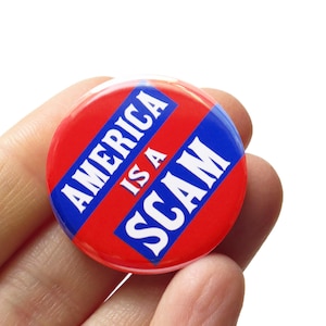 May include: A round button with a red background and the text "AMERICA IS A SCAM" in white letters on a blue banner. The button is held in a hand.