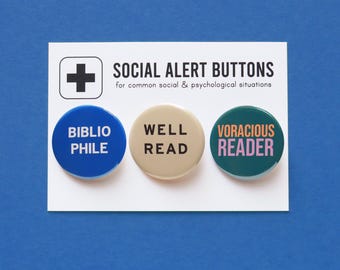 Könnte beinhalten: Drei runde Ansteckbuttons mit weißem Text auf farbigem Hintergrund. Die Buttons zeigen "Bibliophile" auf blauem Hintergrund, "Gut gelesen" auf beigefarbenem Hintergrund und "Vielfraß Leser" auf blaugrünem Hintergrund. Die Buttons sind in einer Reihe auf einer weißen Karte mit dem Text "Social Alert Buttons for common social & psychological situations" angeordnet.
