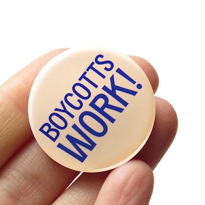 以下が含まれることがあります： 丸い薄いベージュ色のボタンで、太字の濃い青色の大文字で「BOYCOTTS WORK！」と書かれています。白い背景に対して、ボタンは手で持たれています。