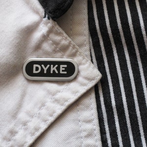 Può includere: Uno spillo d'argento con la parola "DYKE" stampata in lettere nere su sfondo bianco. Lo spillo è attaccato a un tessuto a righe bianche e nere.