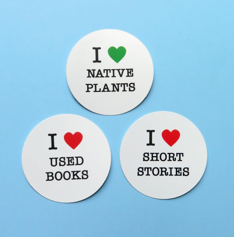 3 round white stickers that read I LOVE NATIVE PLANTS, U LOVE USED BOOKS, I LOVE SHORT STORIES, love being indicated by a red or green heart. Stickers on a light blue background.