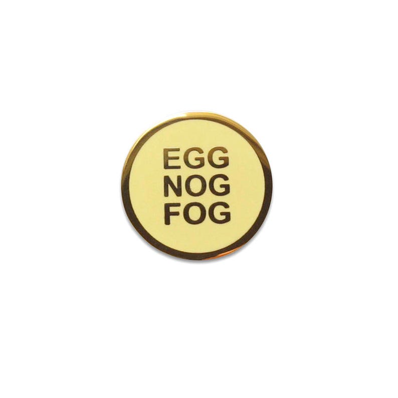 A small round yellowy cream colored enamel pin with a gold outline and three lines of gold text that reads, EGG NOG FOG.