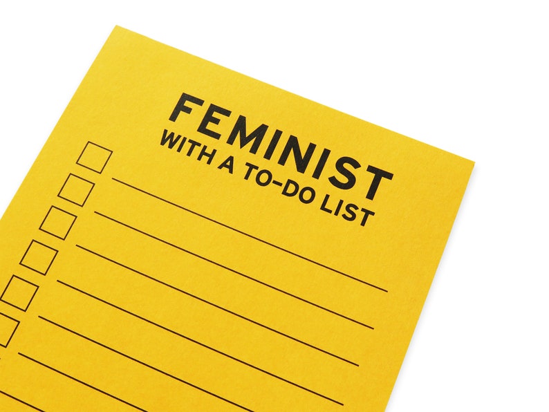 Close up of a vertical rectangle goldenrod notepad that reads FEMINIST WITH A TO-DO LIST at the top, with lines f& check boxes for list making.