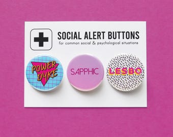 Könnte beinhalten: Drei runde Anstecknadeln mit verschiedenen Designs. Die erste Nadel ist blau mit einem weißen Gittermuster und den Worten "POWER DYKE" in Pink. Die zweite Nadel ist pink mit dem Wort "SAPPHIC" in Weiß. Die dritte Nadel ist weiß mit einem schwarz-weißen geometrischen Muster und dem Wort "LESBO" in Pink und Gelb.