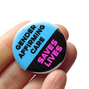 May include: A round button with a light blue and black design. The text "GENDER AFFIRMING CARE" is in dark blue, and "SAVES LIVES" is in pink.