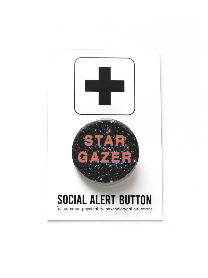 A round black button with a starry space sky background, that reads STAR GAZER in peach & lavender. Badge is on a Social Alert Button backing card.