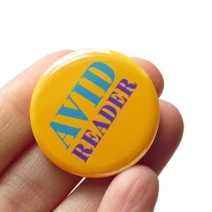 May include: A round, yellow button with the words "AVID READER" in blue and purple. The button is held in a hand against a white background. The button is a pinback button.