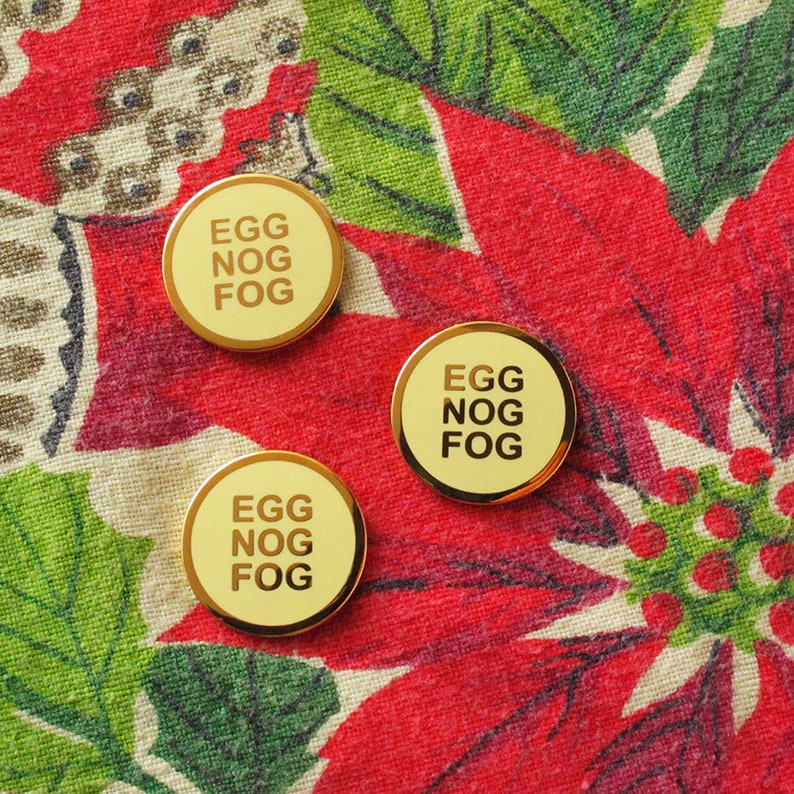 Three small round yellowy cream colored enamel pins with a gold outline and three lines of gold text that reads, EGG NOG FOG. Pinned to a festive red and green Poinsettia tablecloth.