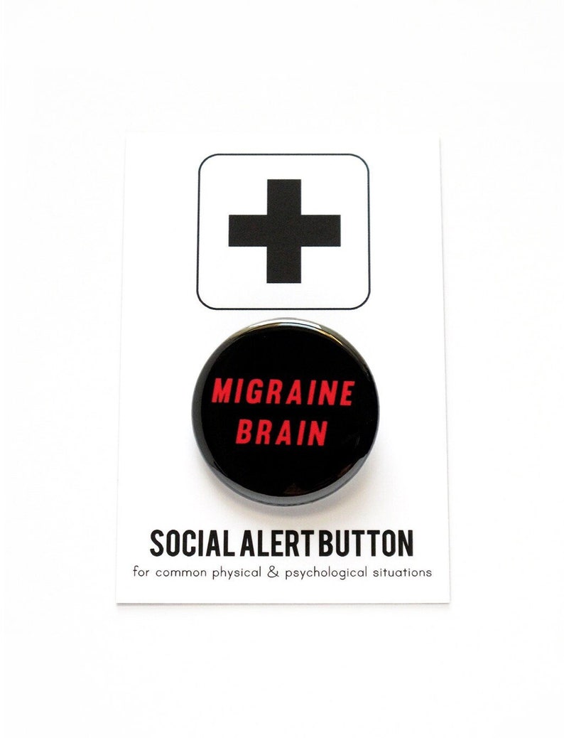 A round black pinback button that reads MIGRAINE BRAIN in red text. Badge is pinned to a Social Alert Button backing card, with a plus sign at the top.