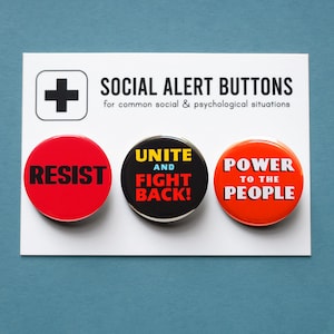 Op de afbeelding: Drie ronde buttons met de teksten: "RESIST" in rood, "UNITE AND FIGHT BACK!" in zwart en "POWER TO THE PEOPLE" in oranje. De buttons liggen op een witte kaart met de tekst "SOCIAL ALERT BUTTONS".