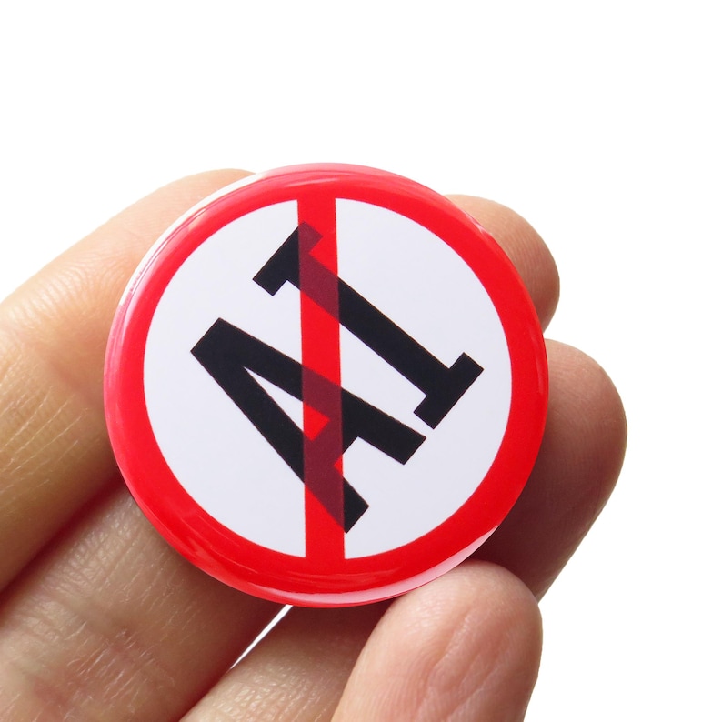 A round white pinback button with a red outline circle-and-slash graphic over the letters 'AI' in black text, indicating the wearer is against AI, or artificial intelligence.  The button is held in a hand.