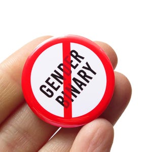 Könnte beinhalten: Ein runder, rot-weißer Button mit den schwarzen Worten "GENDER BINARY", durchgestrichen mit einem roten Strich. Der Button wird in einer Hand gehalten.