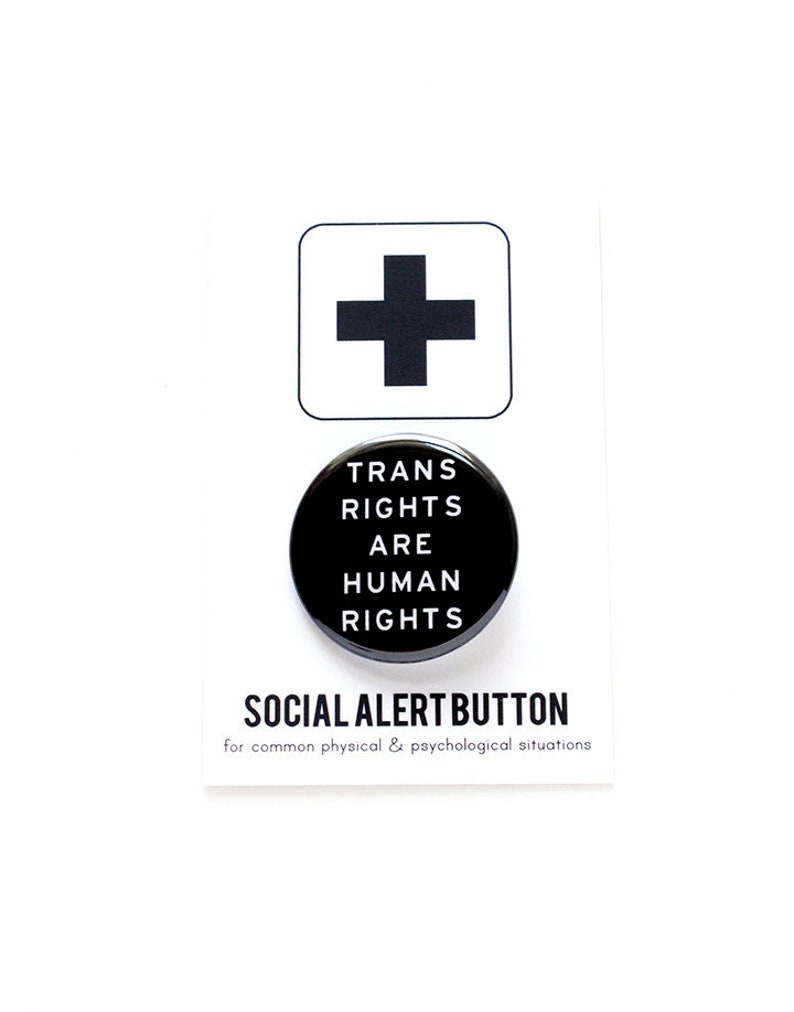 A round black pinback button that reads TRANS RIGHTS ARE HUMAN RIGHTS in white text. Button is on a Social alert Button backing card.