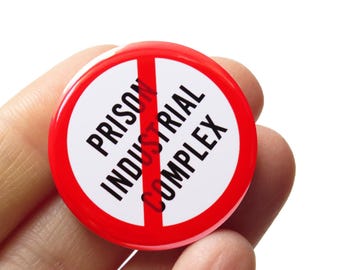 May include: A red and white button with a diagonal red line through it. The text on the button reads "Prison Industrial Complex".