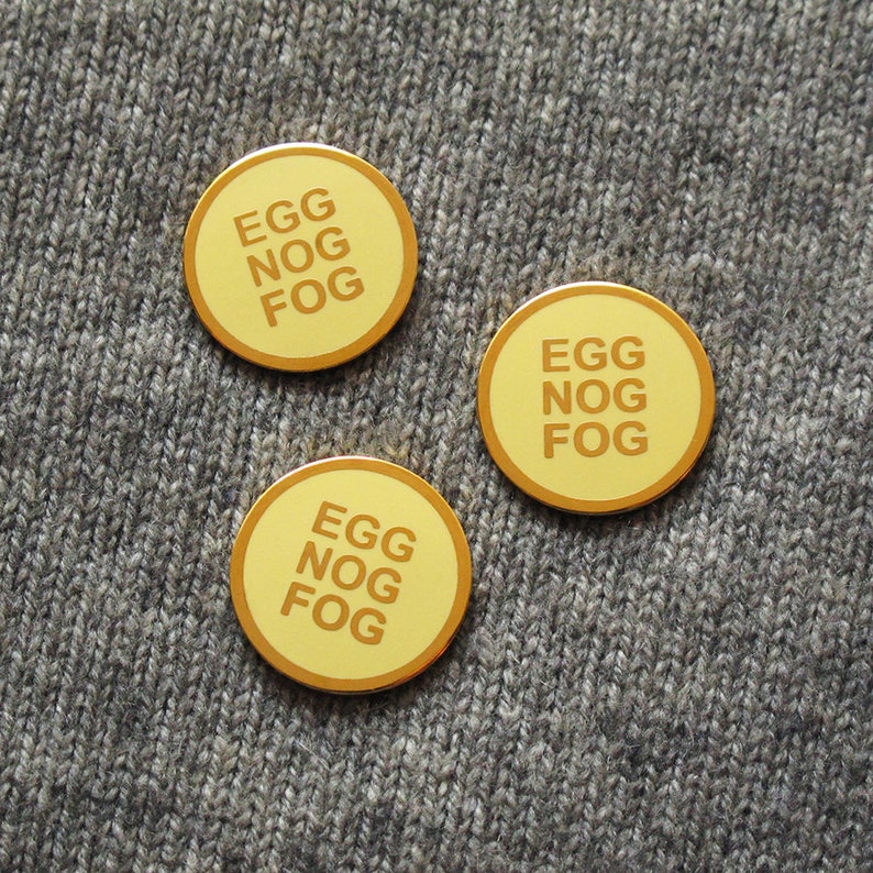 Three small round yellowy cream colored enamel pins with a gold outline and three lines of gold text that reads, EGG NOG FOG. Pinned to a gray sweater