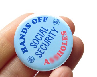 May include: A blue button with the text "Hands Off Social Security A**holes" in white and pink lettering. The button has two dollar signs on it.