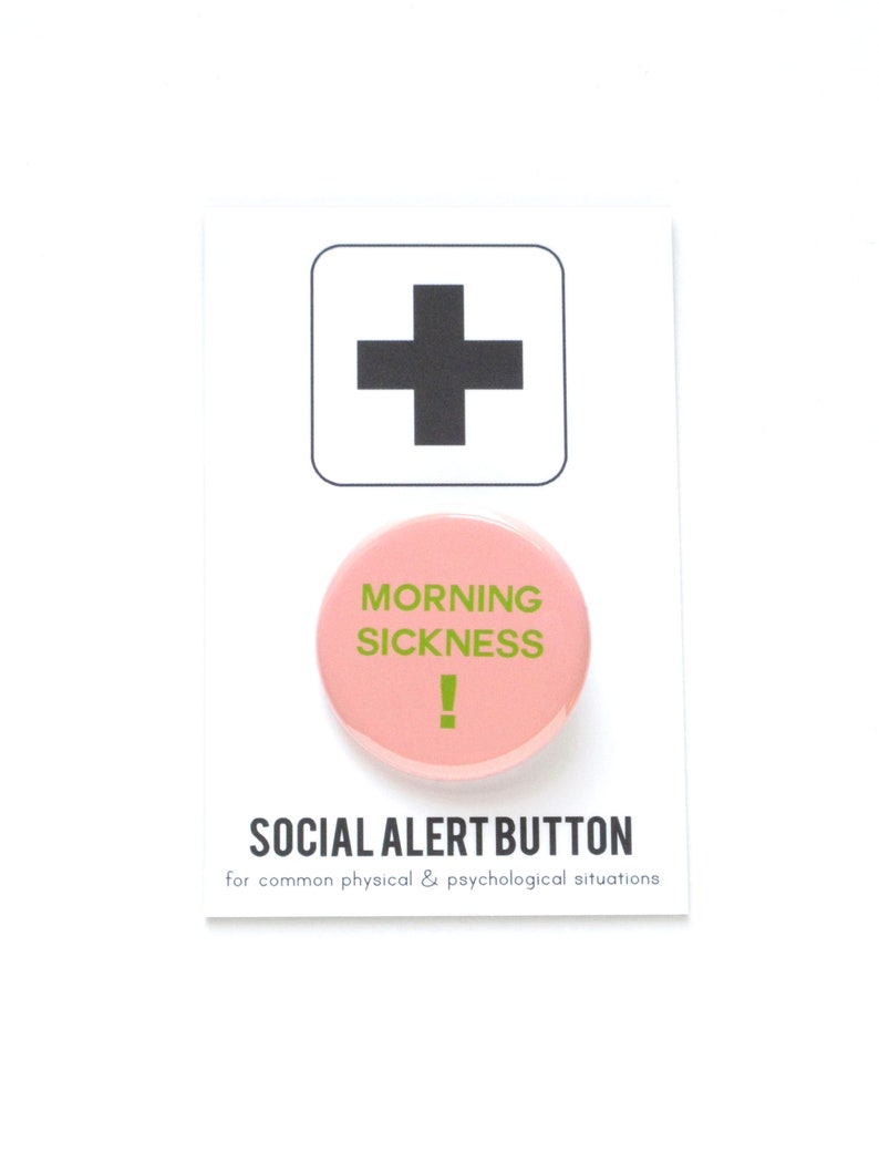 A round light pink pinback button that reads MORNING SICKNESS ! in puke green text. Pin is on a Social Alert Button backing card.