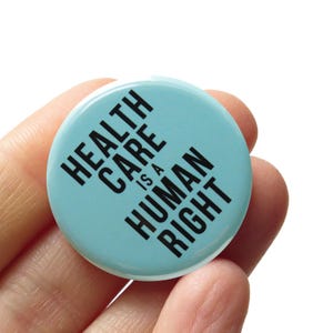 Può includere: Un bottone azzurro chiaro con il testo "HEALTH CARE IS A HUMAN RIGHT" in nero.