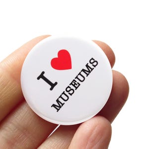 May include: A circular white pin-back button featuring a red heart symbol followed by the black text I MUSEUMS. The design is printed on a smooth, glossy surface with a clean, minimalist aesthetic, clearly displaying the message in a bold font.