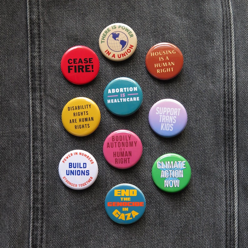 Multi-color social justice pinback buttons on black denim, with messages such as: Abortion Is Healthcare, Disability Rights Are Human Rights, Housing Is A Human Right, There is Power In A Union, Support Trans Kids, Climate Action Now, Ceasefire etc.