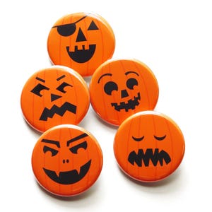May include: Five orange pumpkin-shaped buttons with different black jack-o'-lantern faces. The faces include a pirate, a surprised face, a frowning face, a vampire, and a sleepy face.
