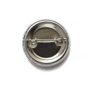 May include: The back of a round, silver button with a white border. The button has a metal pin on the back for attaching to clothing.