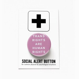May include: A pink button with the text "TRANS RIGHTS ARE HUMAN RIGHTS" in white. The button is on a white card with a black plus sign and the text "SOCIAL ALERT BUTTON for common physical & psychological situations".