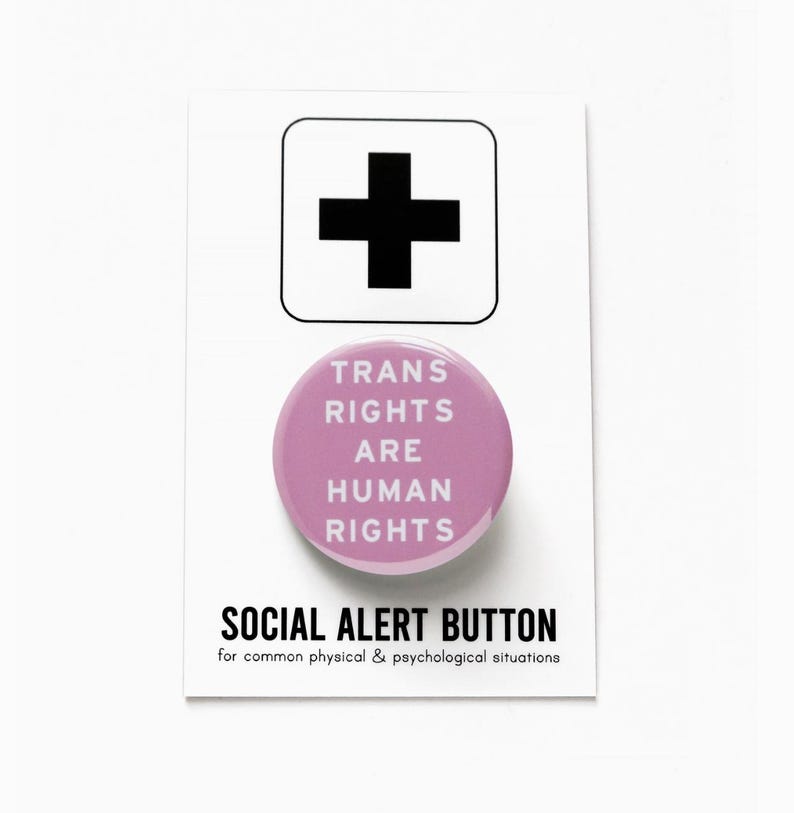May include: A pink button with the text "TRANS RIGHTS ARE HUMAN RIGHTS" in white. The button is on a white card with a black plus sign and the text "SOCIAL ALERT BUTTON for common physical & psychological situations".