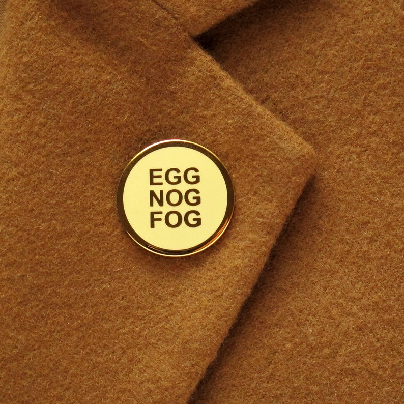 A small round yellowy cream colored enamel pin with a gold outline and three lines of gold text that reads, EGG NOG FOG. Pinned to a soft camel colored jacket lapel.